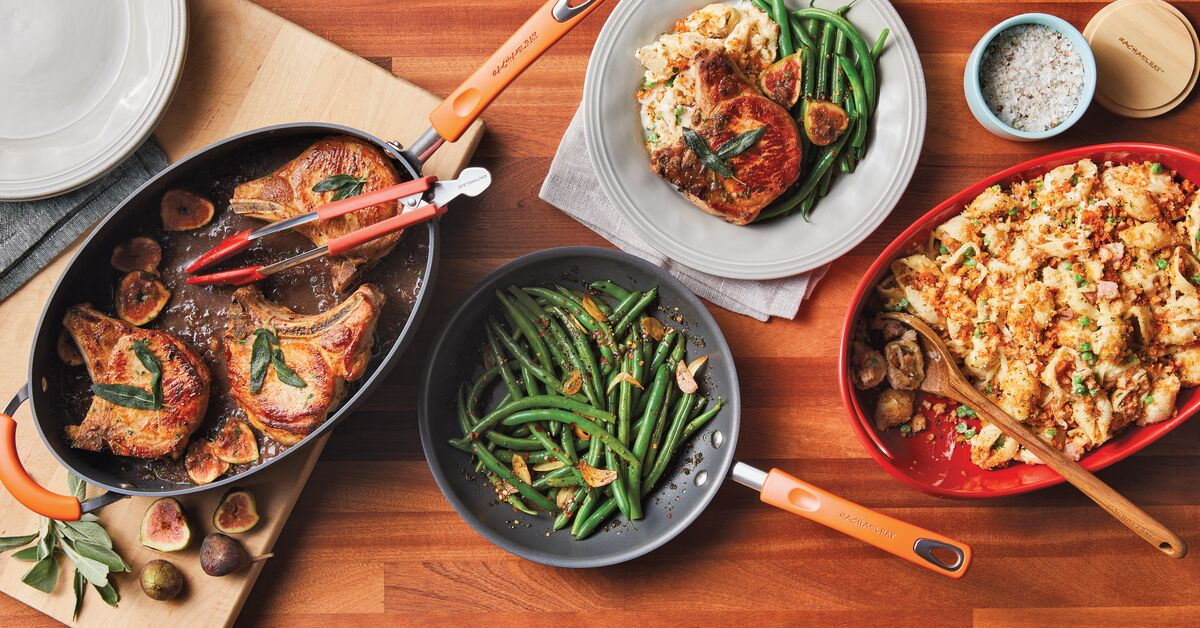 Two pans with green beans and pork chops, a plate with pork chops and beans, and a red tray of pasta mixed with vegetables on a wooden table.