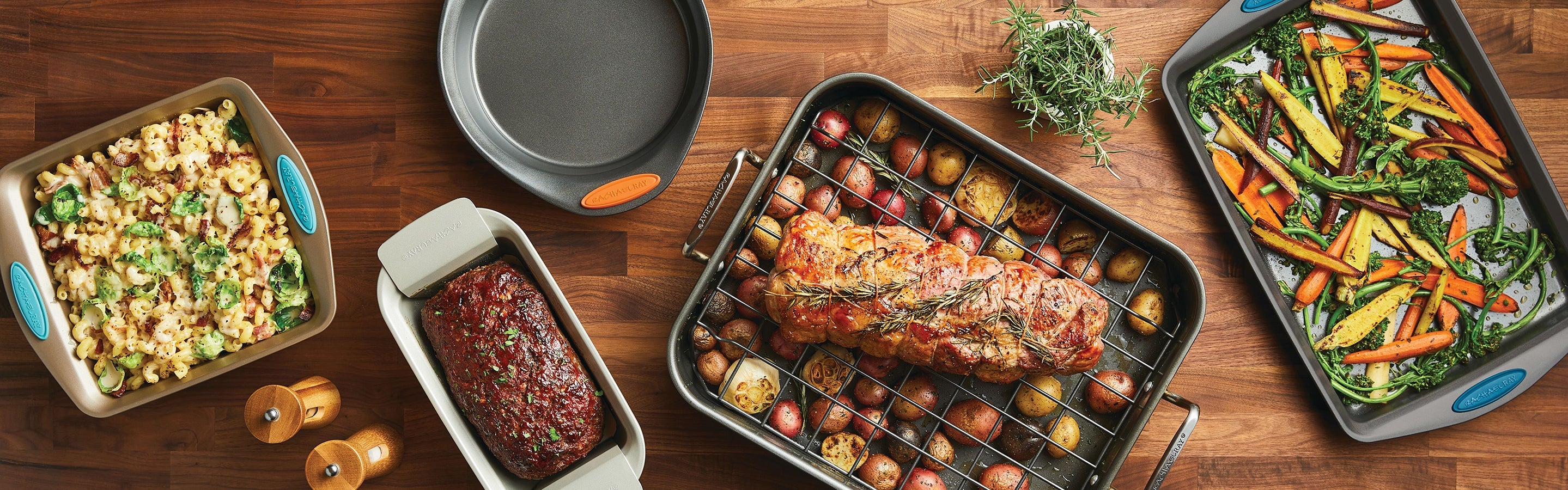 A wooden table displays baked macaroni, meatloaf, a roast with potatoes, a round cake pan, and roasted vegetables in colorful bakeware.