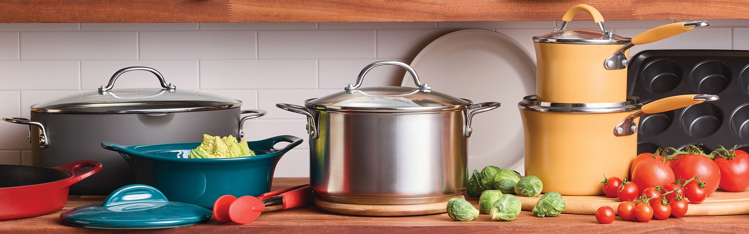 Various pots, pans, fresh vegetables, and kitchen utensils are arranged neatly on a wooden countertop in front of a white tiled backsplash.
