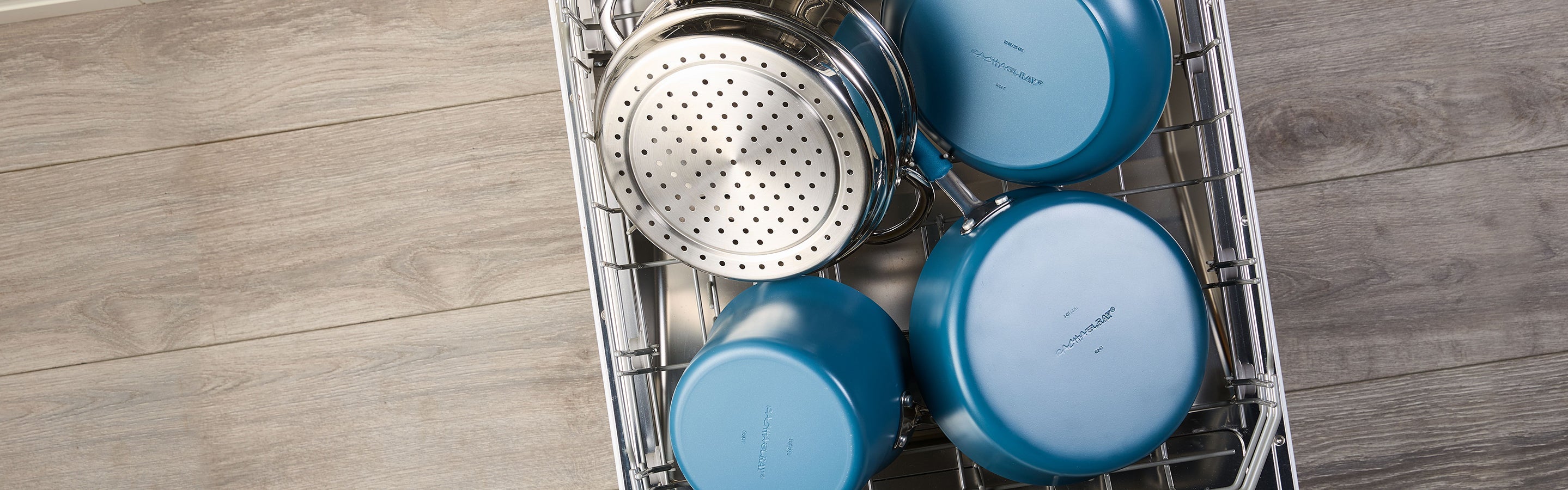 Four blue nonstick pots and pans and a metal strainer are arranged in the top rack of an open dishwasher on a wood floor.