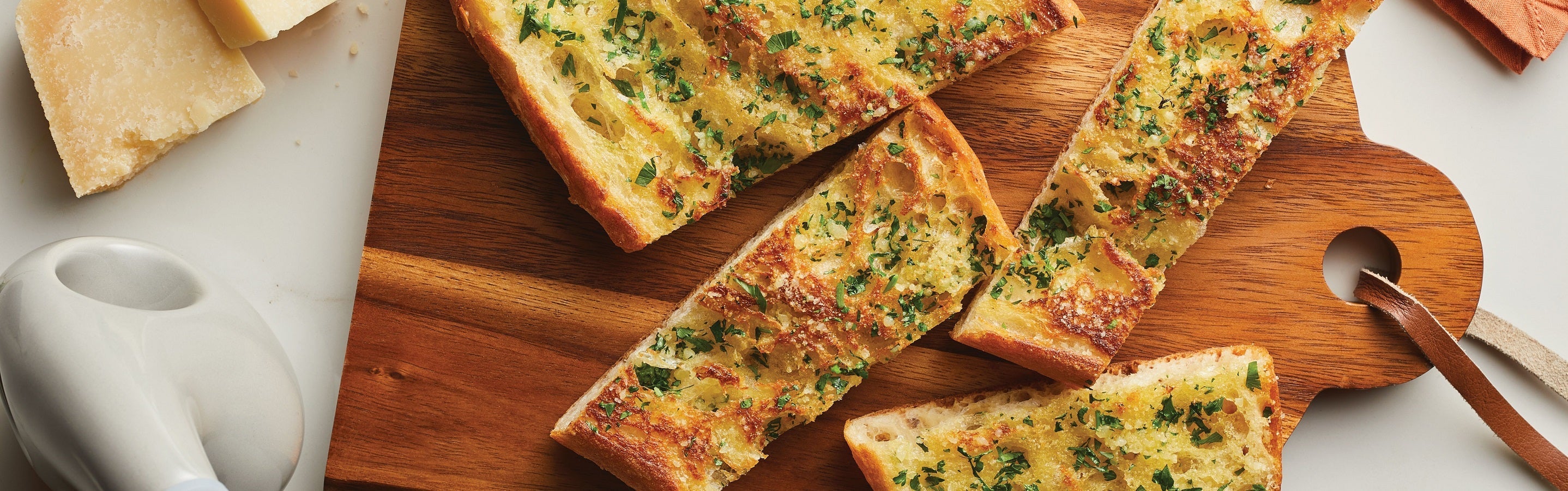 Sliced garlic bread with herbs on a wooden cutting board, accompanied by a cheese wedge and a white ceramic dish.