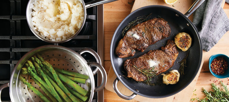 Steak with butter and garlic in a pan, mashed potatoes in a pot, and asparagus in a steamer basket on a stovetop.