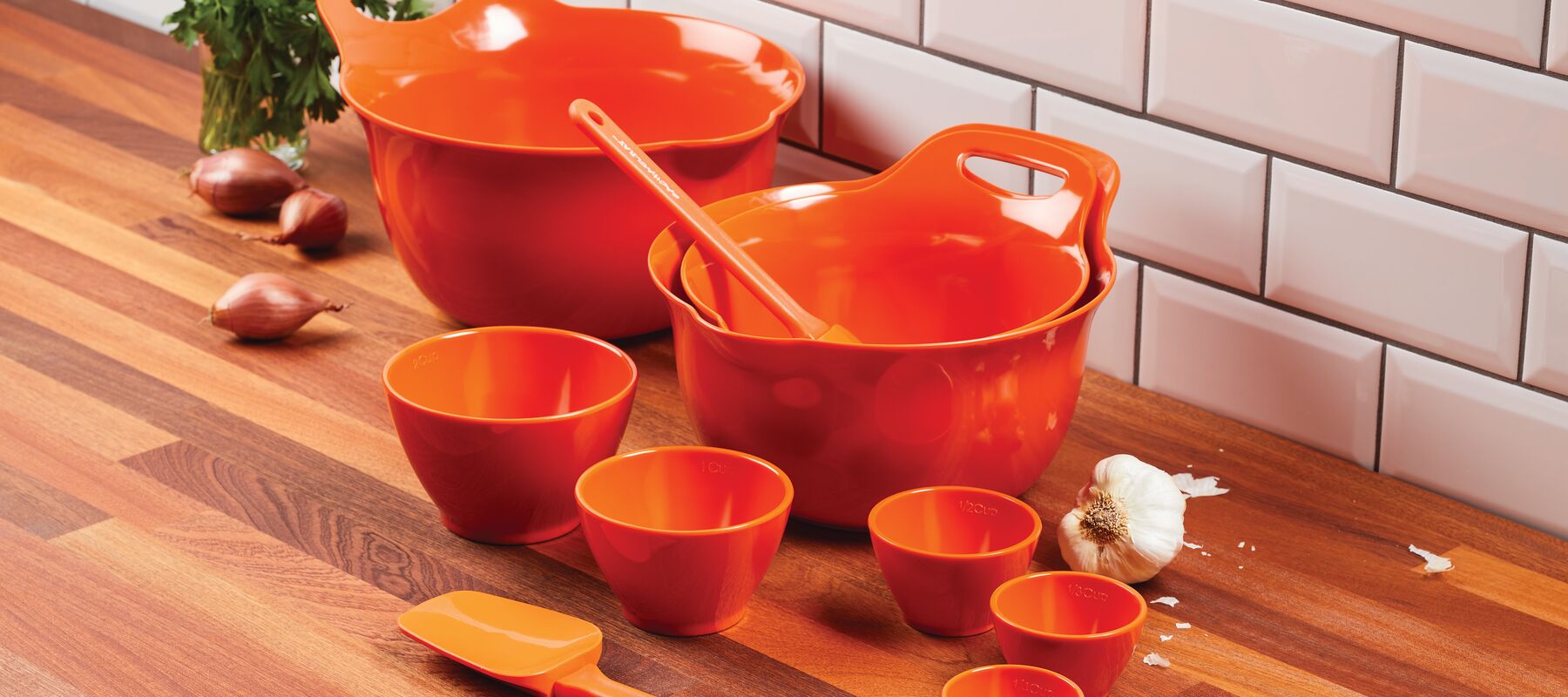 Bright orange mixing bowls, measuring cups, and spatulas are arranged on a wooden kitchen counter with onions and garlic nearby. White subway tiles form the backsplash.