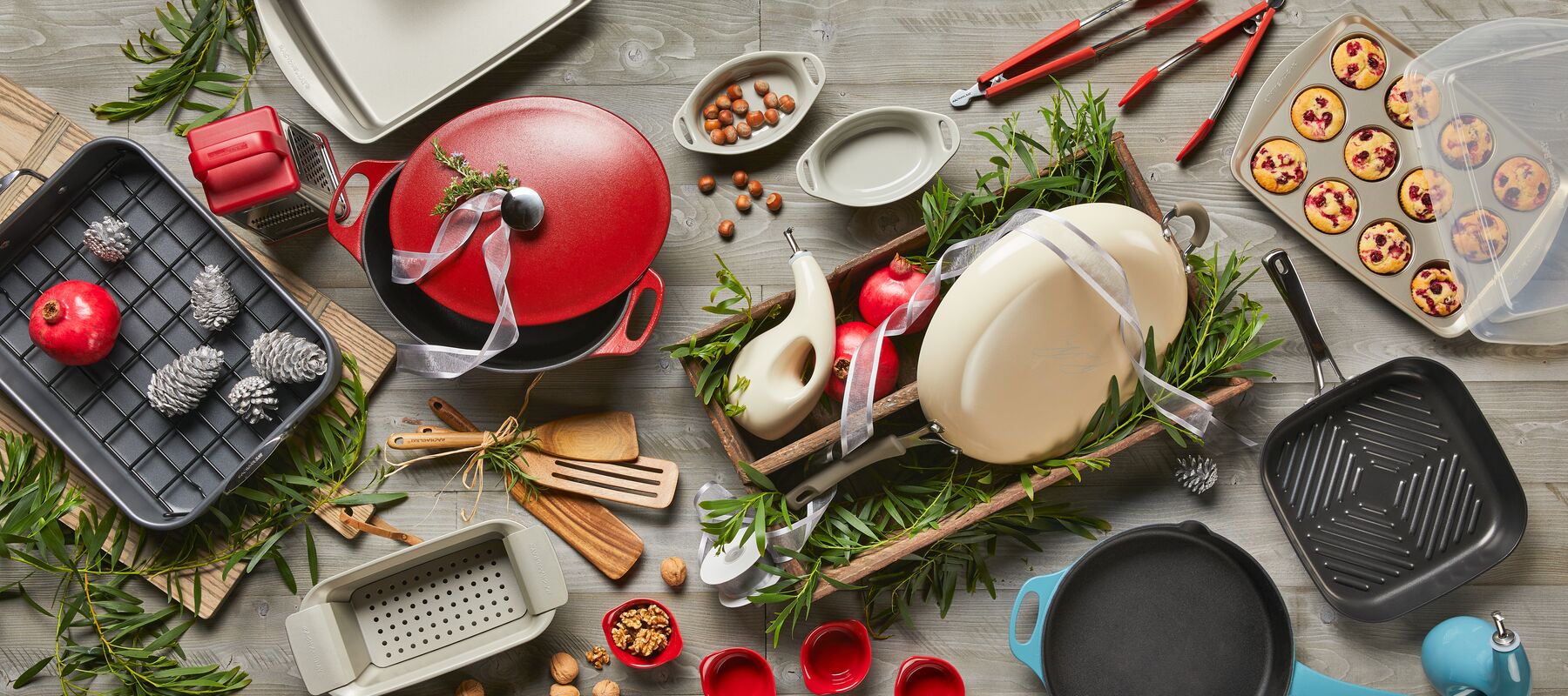 A variety of kitchenware is arranged on a wooden surface, including pans, a red pot, baking trays with pastries, utensils, pomegranates, rosemary, and pine cones.