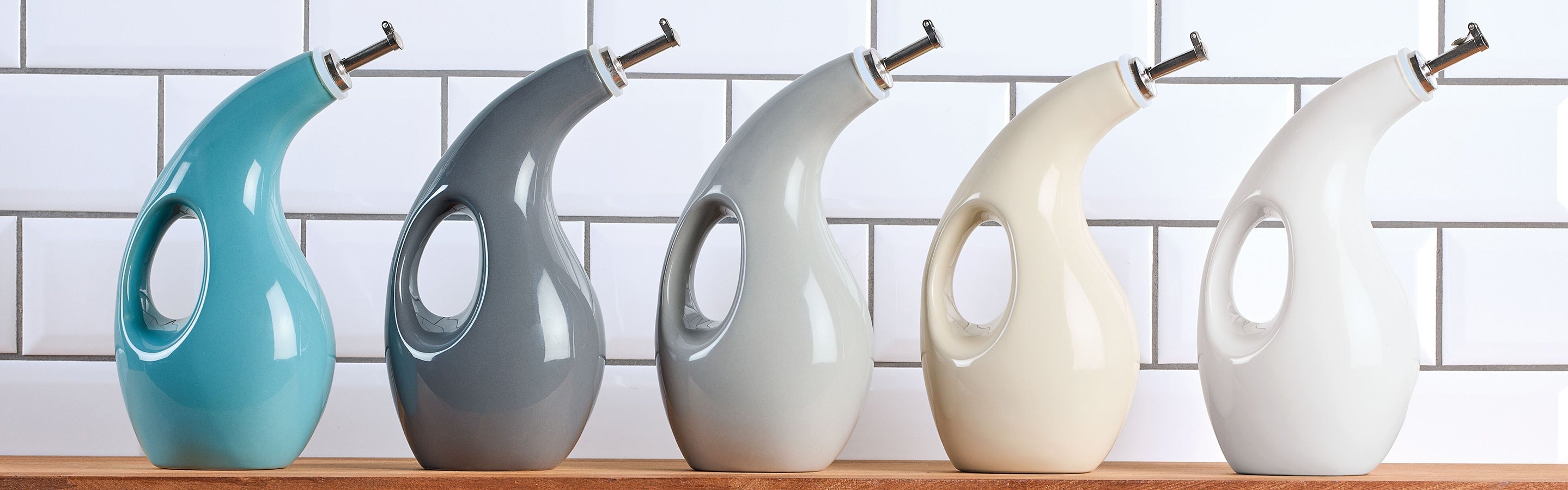 Five ceramic oil dispensers in different colors are arranged in a row on a wooden shelf against a white tiled wall.