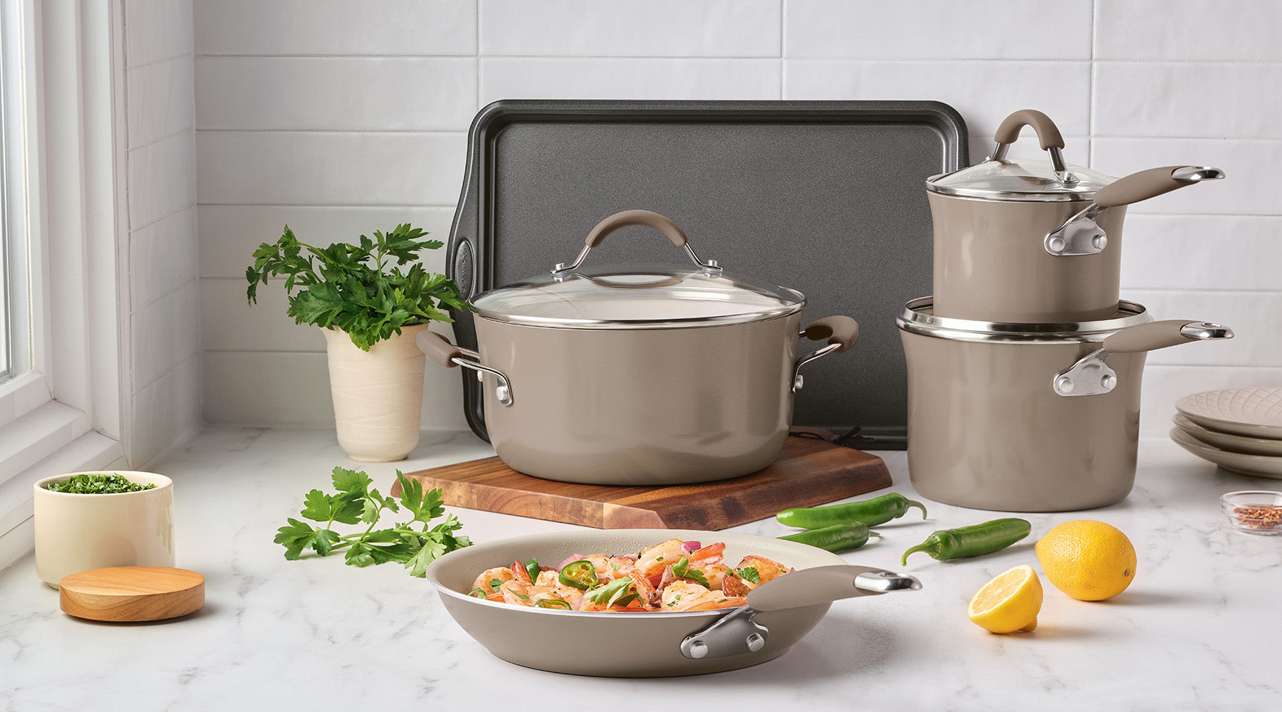 A set of beige cookware with lids displayed on a kitchen counter, surrounded by herbs, lemons, a baking tray, plates, and a prepared vegetable dish.