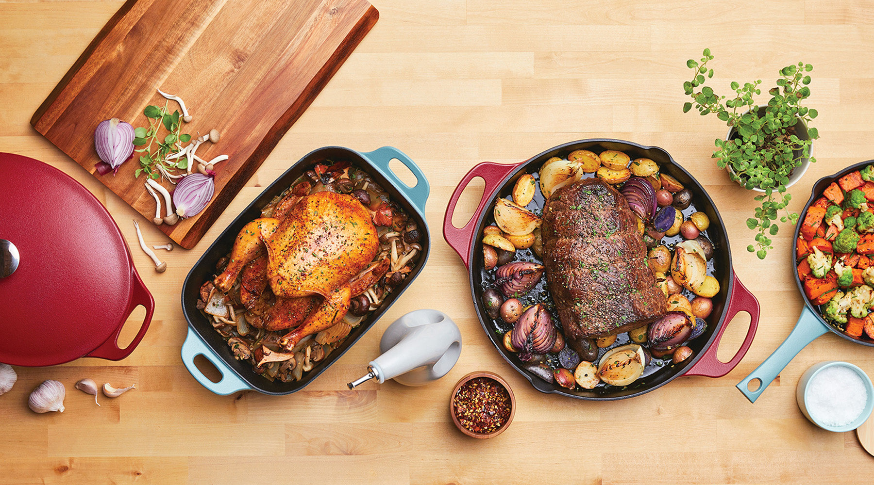 Three cast iron dishes on a wooden table: one with roast chicken, one with roast beef and potatoes, and one with roasted vegetables; various herbs and ingredients around them.