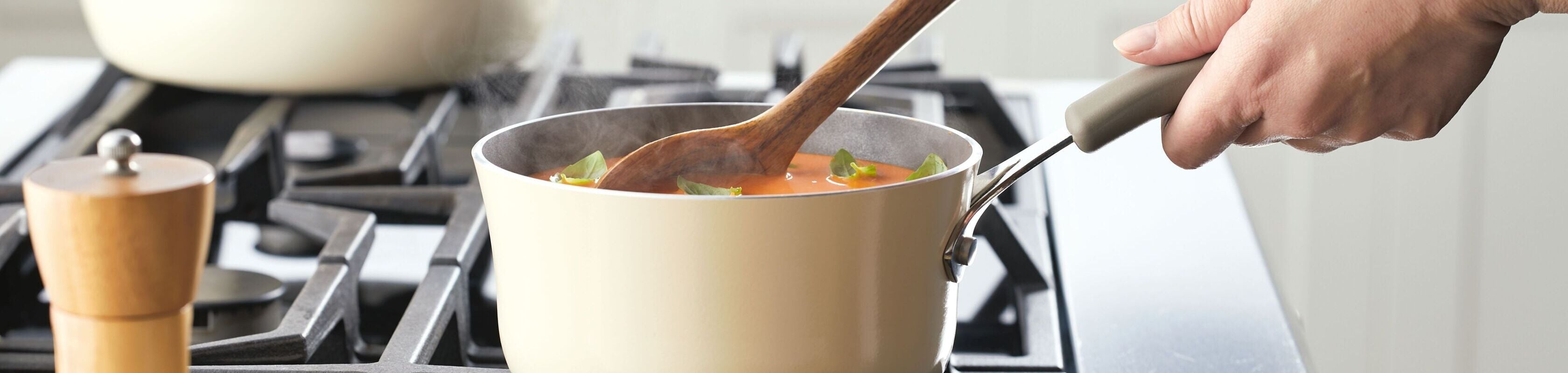 A person stirs a pot of soup with a wooden spoon on a stovetop; vegetables are visible in the pot and a pepper grinder sits nearby.