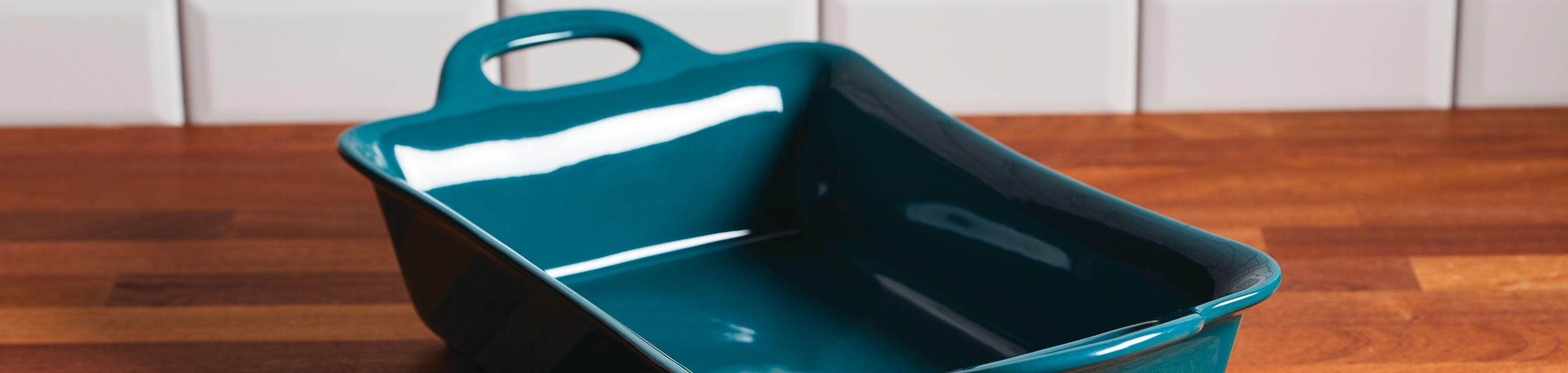A rectangular, deep teal ceramic baking dish with handles sits on a wooden countertop in front of white tile backsplash.