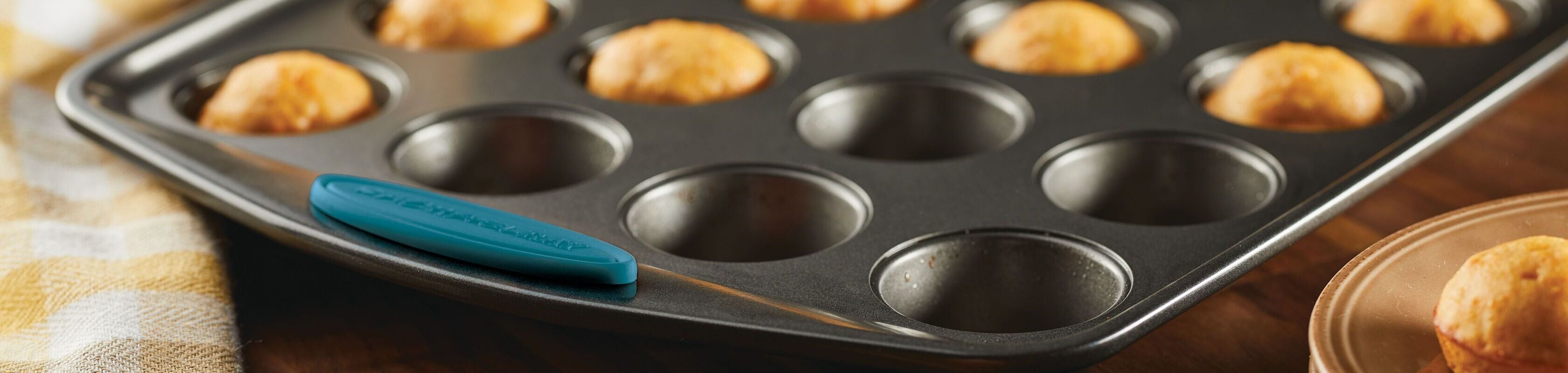 A nonstick muffin tin with several unbaked dough balls sits on a wooden surface next to a checkered cloth.