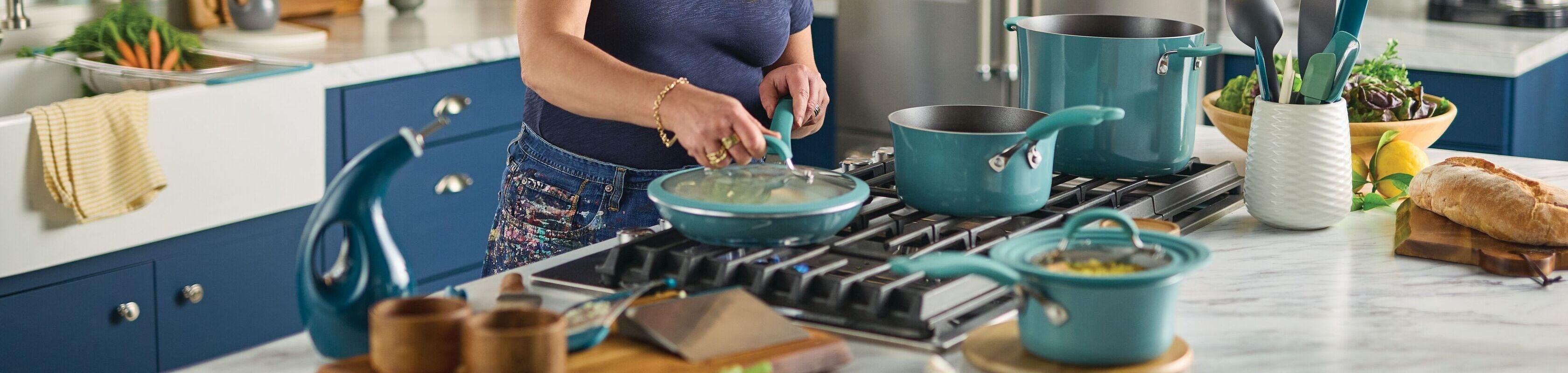 A woman cooks at a modern kitchen stove with blue cookware, surrounded by fresh ingredients, utensils, and bread on the counter.