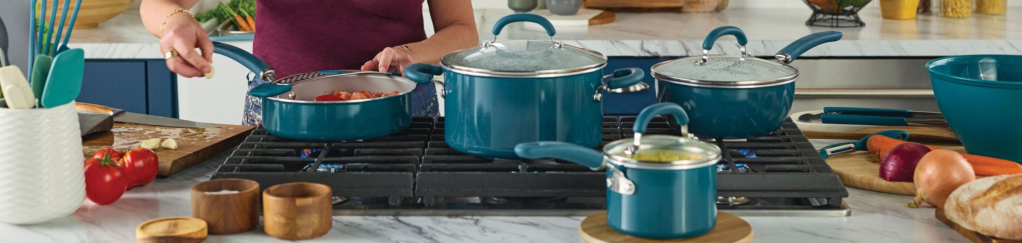 A woman cooks at a modern kitchen stove with blue cookware, surrounded by fresh ingredients, utensils, and bread on the counter.