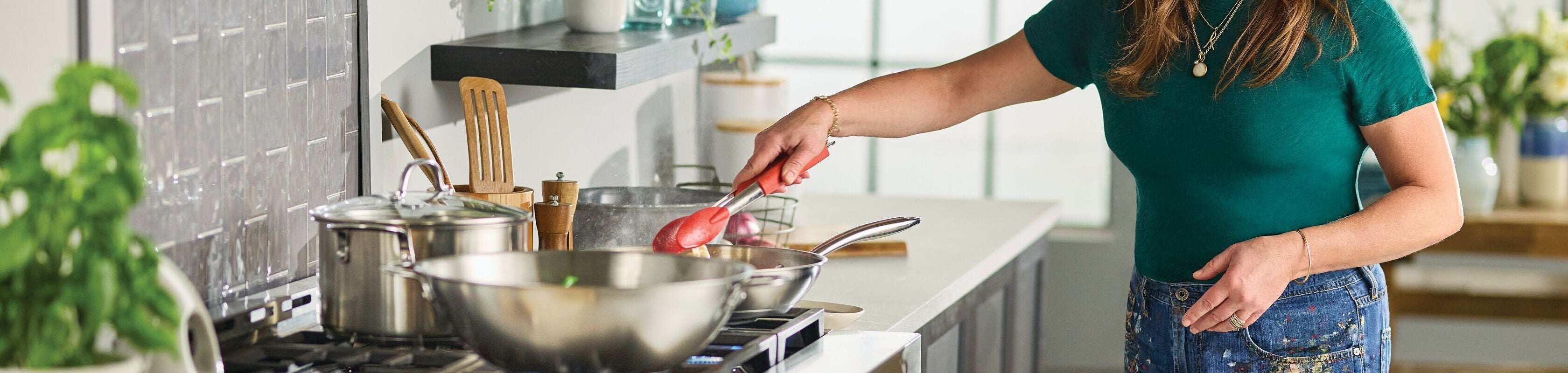 A woman in a green shirt uses red tongs to cook at a stovetop in a modern kitchen with pots, pans, and utensils visible.