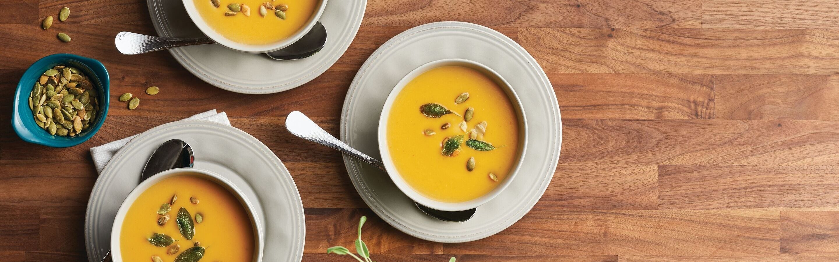Three bowls of orange soup topped with seeds, each on a plate with a spoon, sit on a wooden surface next to a small bowl of pumpkin seeds.