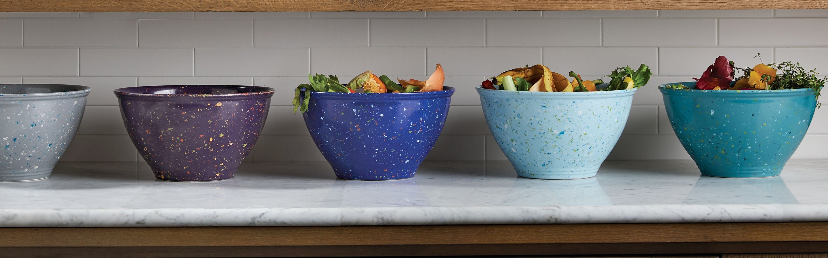 Five ceramic bowls in varying shades of blue and purple are lined up on a marble countertop, each filled with assorted vegetable scraps.