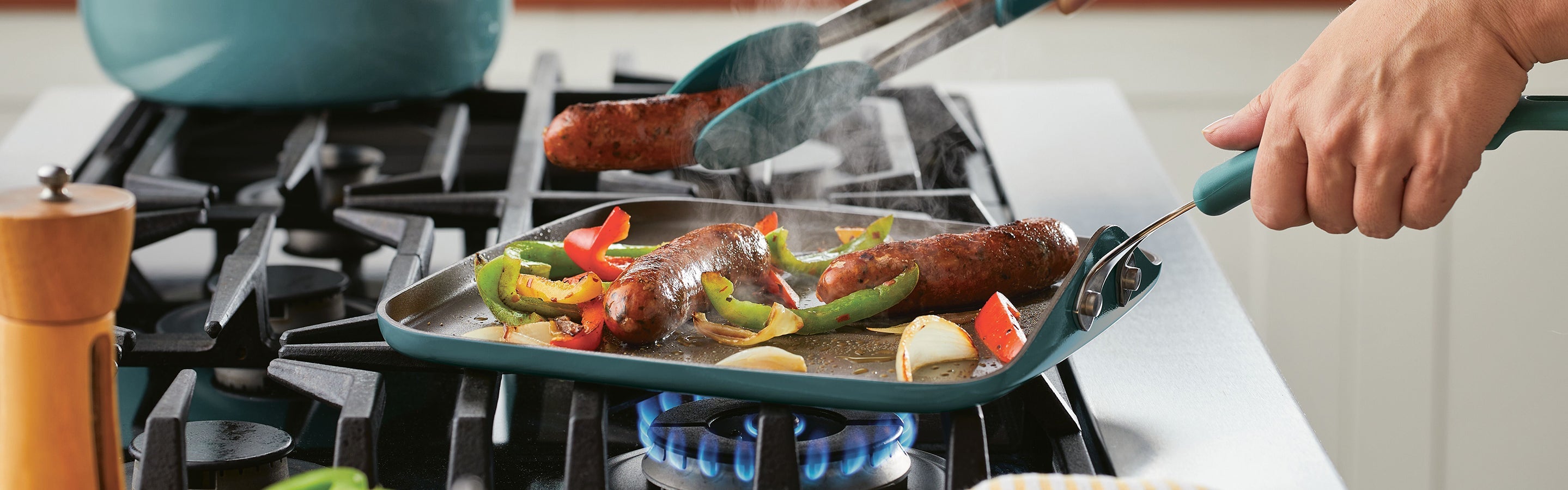Sizzling sausages and sliced peppers being cooked on a grill pan over a gas stove, with tongs lifting one sausage.