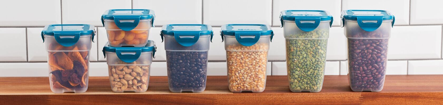 Four plastic food storage containers with teal lids are stacked on a wooden countertop, each filled with different foods like oranges, salad, and vegetables. White tile wall in background.