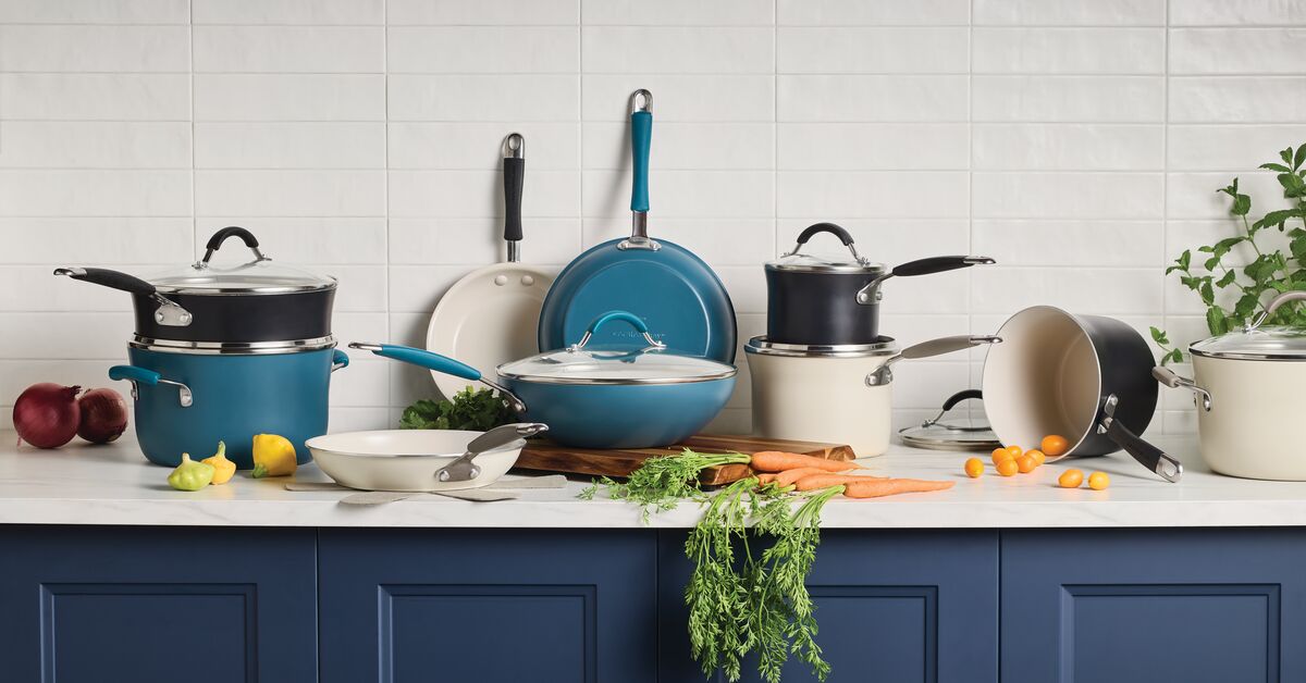 A variety of cookware, including pots and pans, displayed on a kitchen counter with vegetables, against a white tile backsplash.