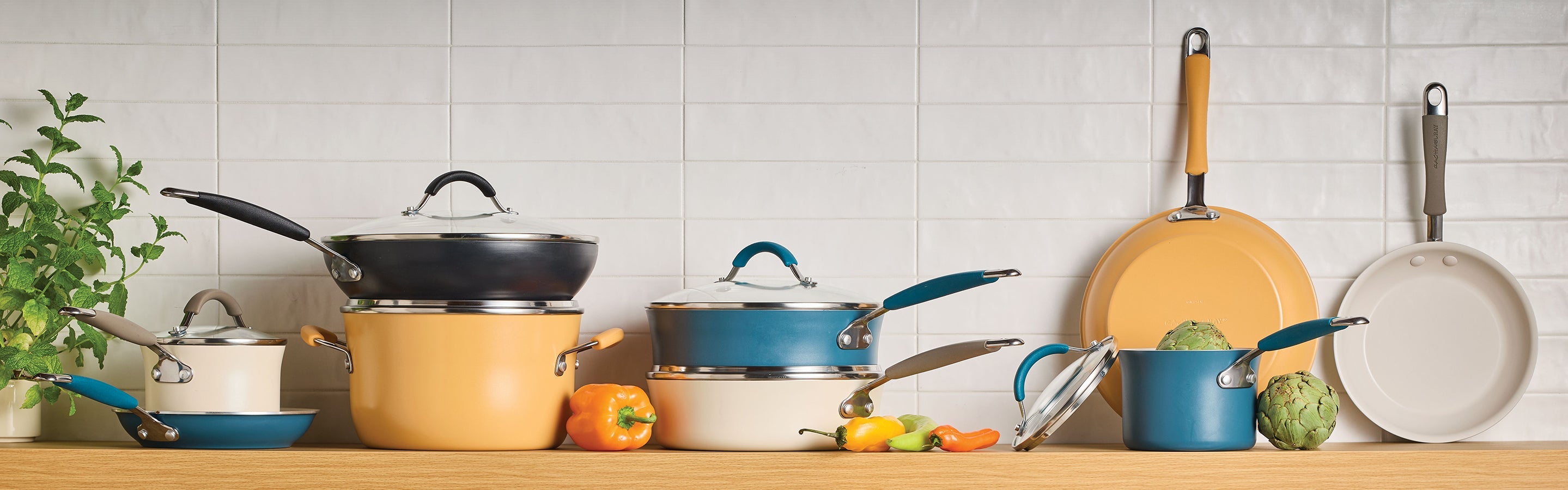 A variety of cookware, including pots and pans, displayed on a kitchen counter with vegetables, against a white tile backsplash.