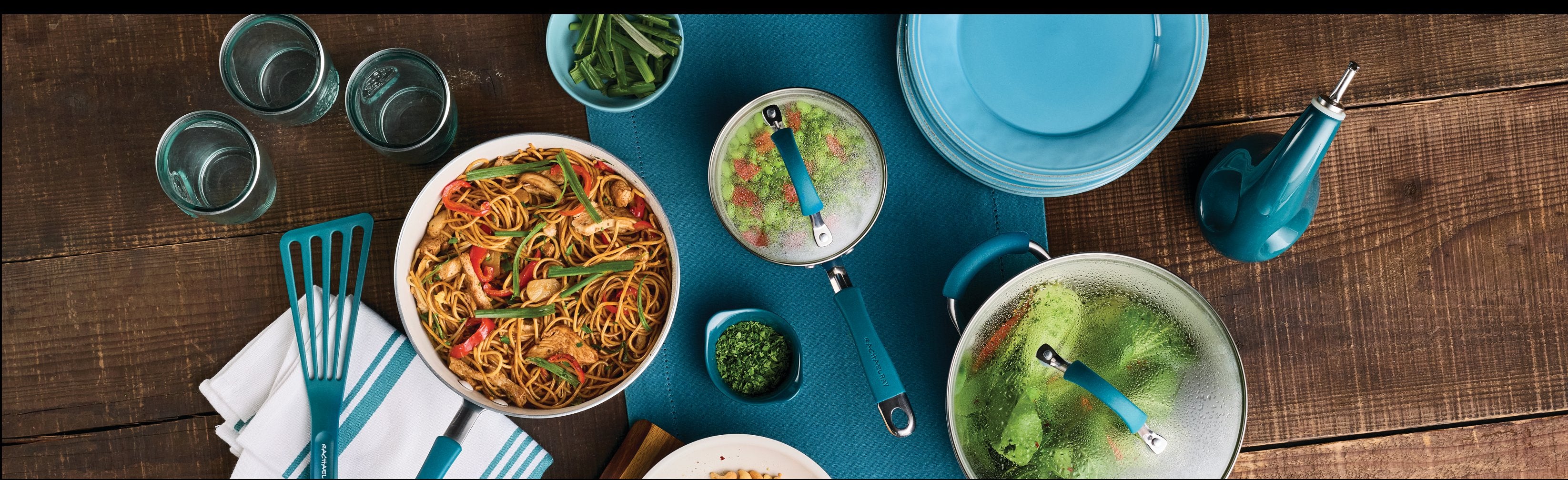 A collection of cookware, including pots and pans with lids, displayed on a countertop with vegetables. Navy and white cabinets are visible below, with a tiled wall in the background.