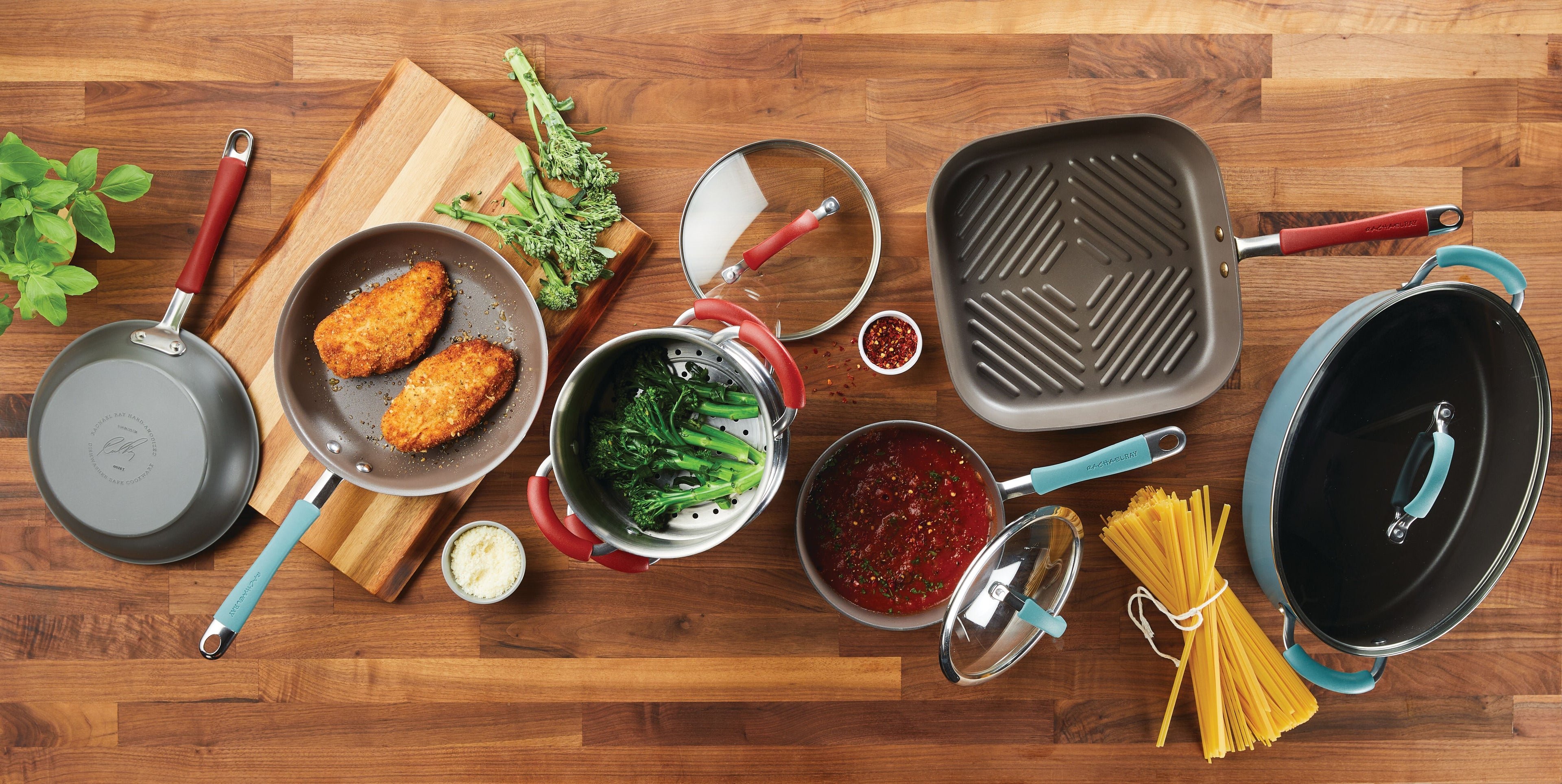 Assorted cookware on a wooden table with cooked chicken, pasta, broccoli, sauce, and a grill pan, surrounded by lids and utensils.