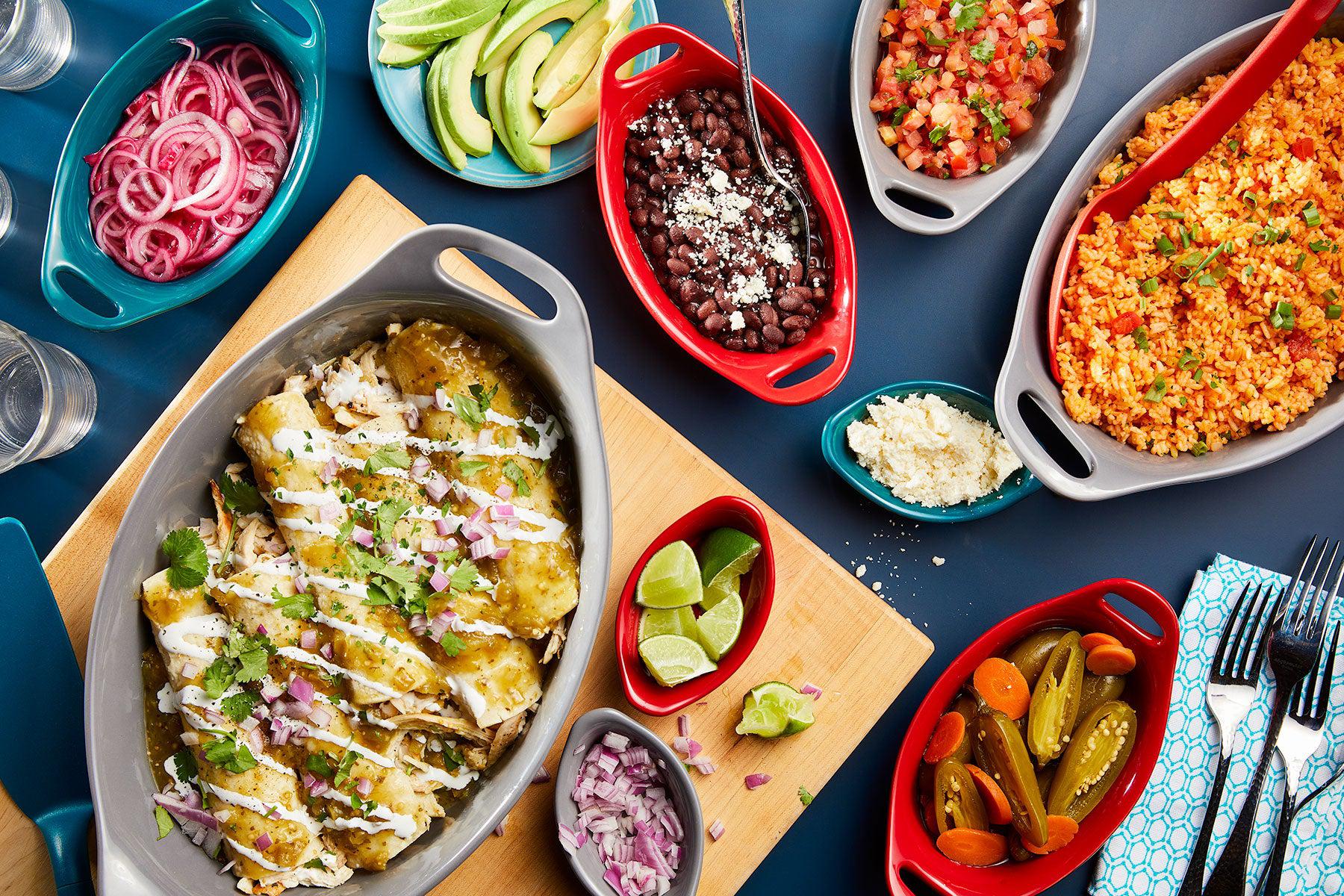 A table set with enchiladas, Mexican rice, black beans, pickled onions, sliced avocados, pico de gallo, limes, pickled vegetables, and garnishes on colorful dishes.