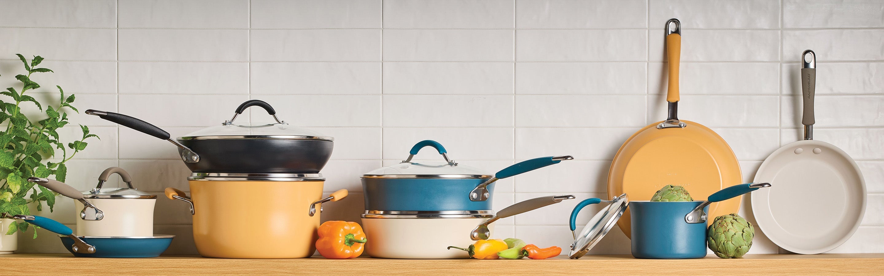 Various pots, pans, and lids in cream, blue, and yellow colors are arranged on a kitchen counter in front of a white tile wall, with vegetables and a plant nearby.