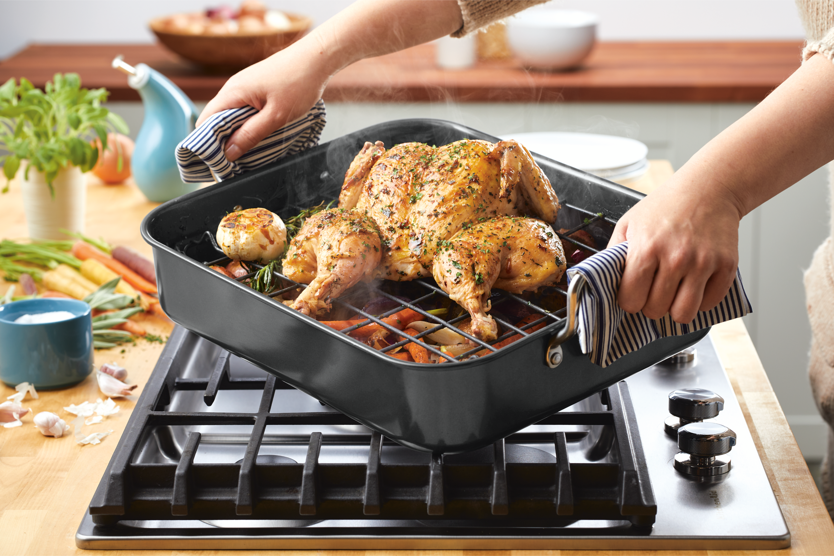 A person places a roasted chicken in a black pan with vegetables onto a stovetop, using striped oven mitts for protection.