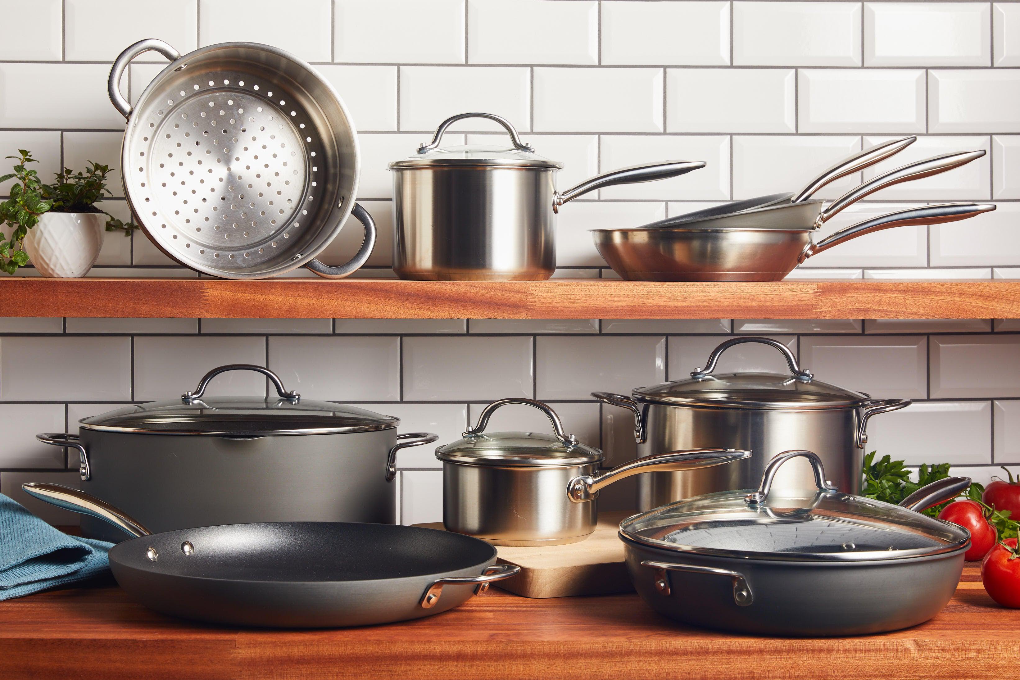 A collection of stainless steel and non-stick cookware including pots, pans, and a colander, displayed on wooden shelves against a white tiled backsplash.