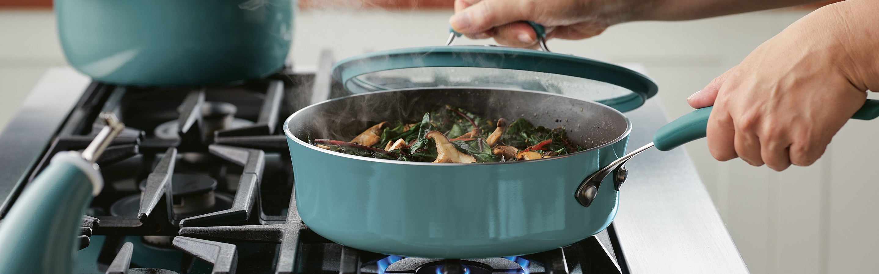 A person lifts the lid off a teal saucepan on a gas stove, revealing steaming vegetables and mushrooms cooking inside.