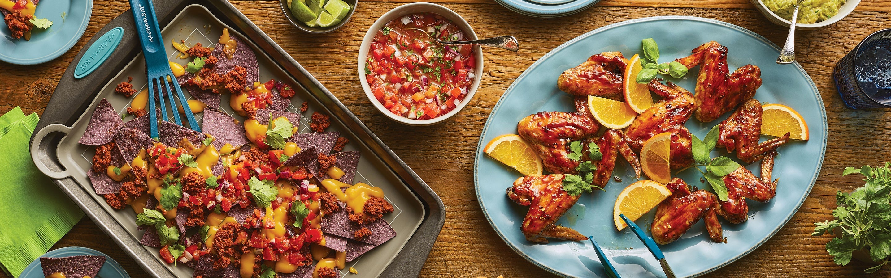 A tray of blue corn nachos with cheese and salsa is next to a plate of BBQ chicken wings garnished with orange slices and herbs; a bowl of salsa sits nearby.