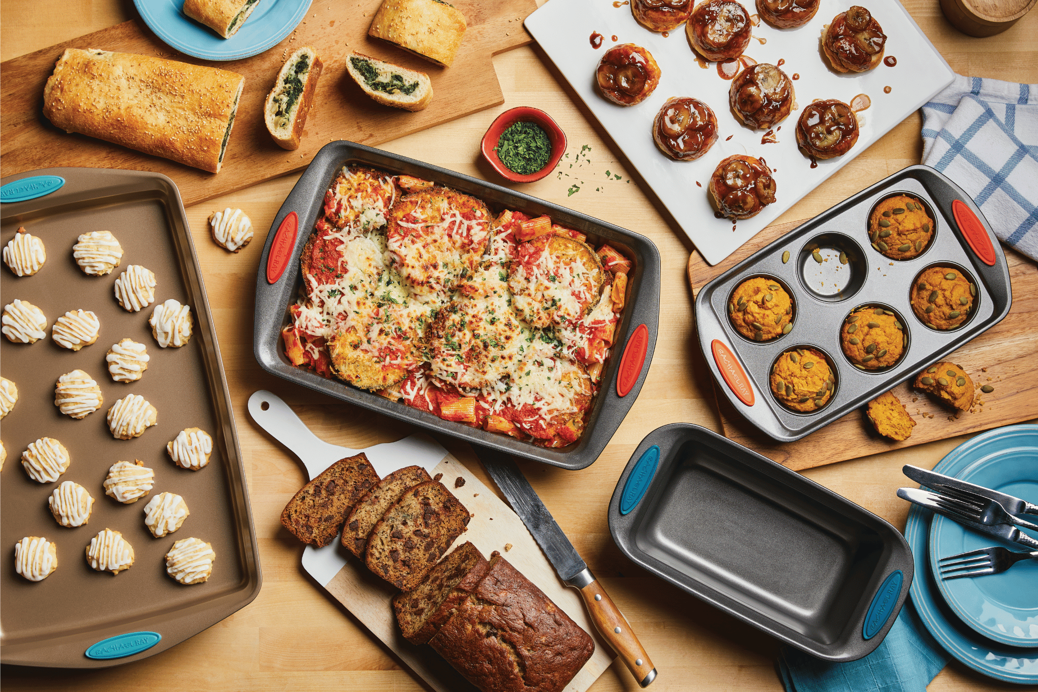 Assorted baked dishes on a wooden table, including lasagna, muffins, cookies, bread, stuffed pastry, and meatballs, surrounded by baking pans and serving plates.