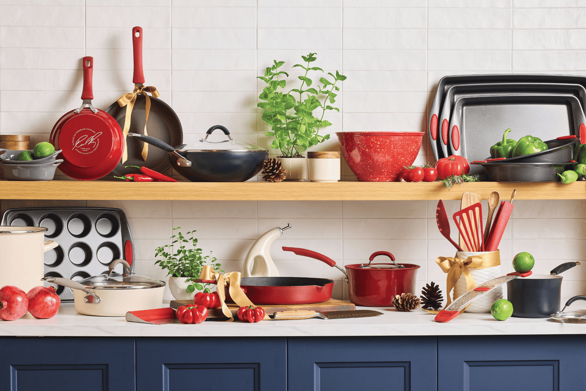 A kitchen counter and shelves display assorted red cookware, utensils, bakeware, vegetables, herbs, and decorative items against a white tile wall.