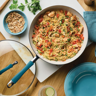 Pan of pasta with shrimp and vegetables on a wooden table, with plates nearby.