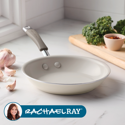 Ceramic frying pan on counter with garlic, greens, and spices nearby.