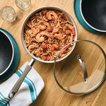 Pan of shrimp pasta on a wooden table with plates and glasses nearby.