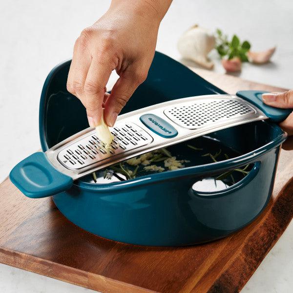 Using the Stainless Steel Multi-Grater, grate garlic into a blue bowl while enjoying the comfortable grip provided by its silicone handles.