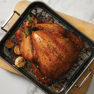 A roasted turkey sits in the 16-Inch x 12-Inch Nonstick Hard Anodized Roaster with Reversible Rack on a wooden cutting board, offering effortless cleanup and consistent cooking results.