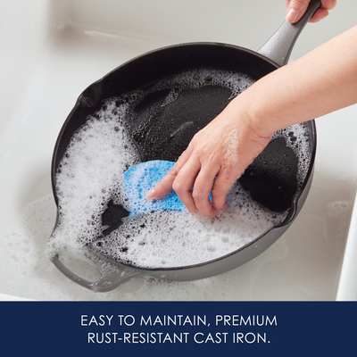 Person washing a cast iron skillet in soapy water with a blue sponge.