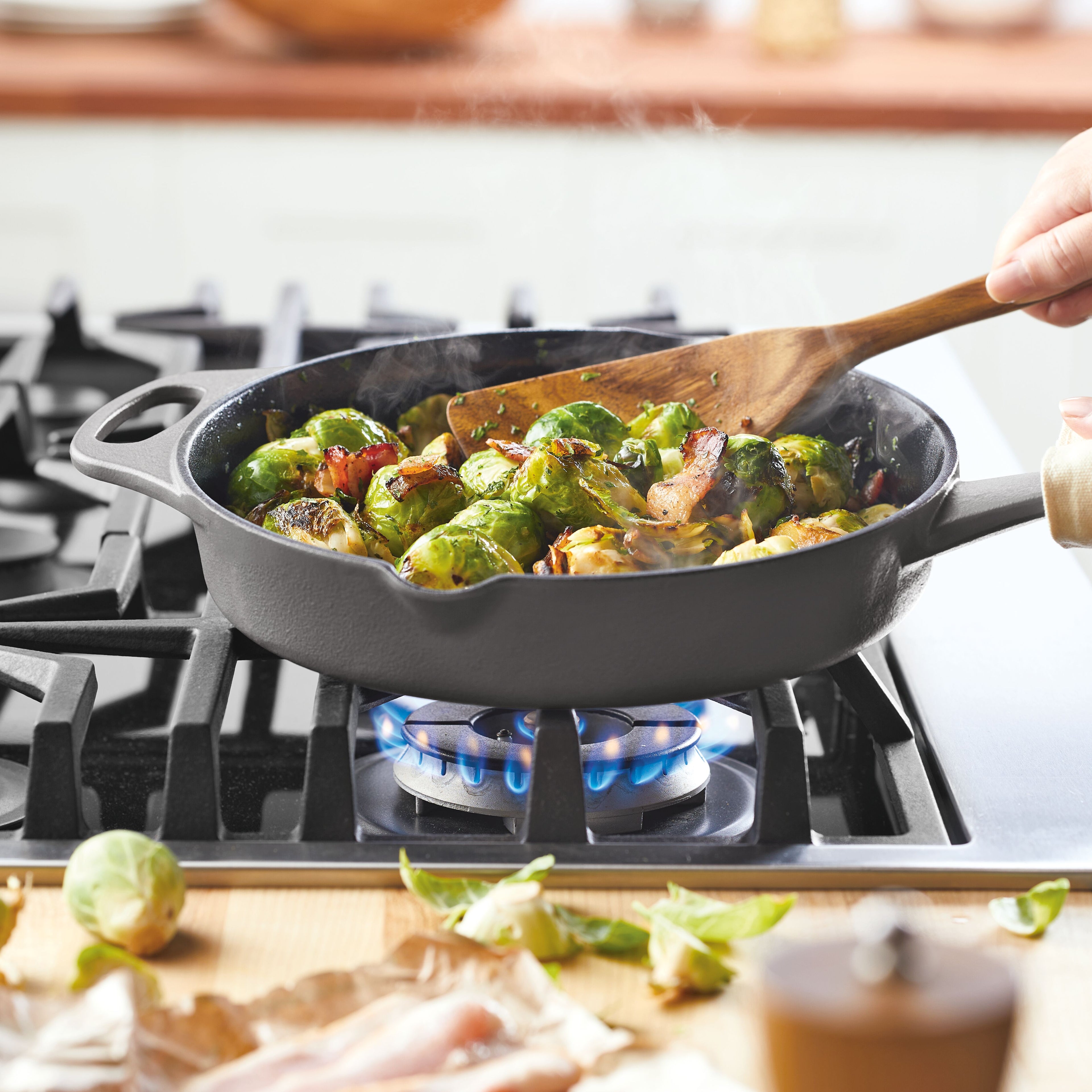 A close-up of a gray Rachael Ray NITRO Cast Iron 10-Inch Frying Pan on a gas stovetop, featuring roasted Brussels sprouts and bacon being stirred with a wooden spatula.
