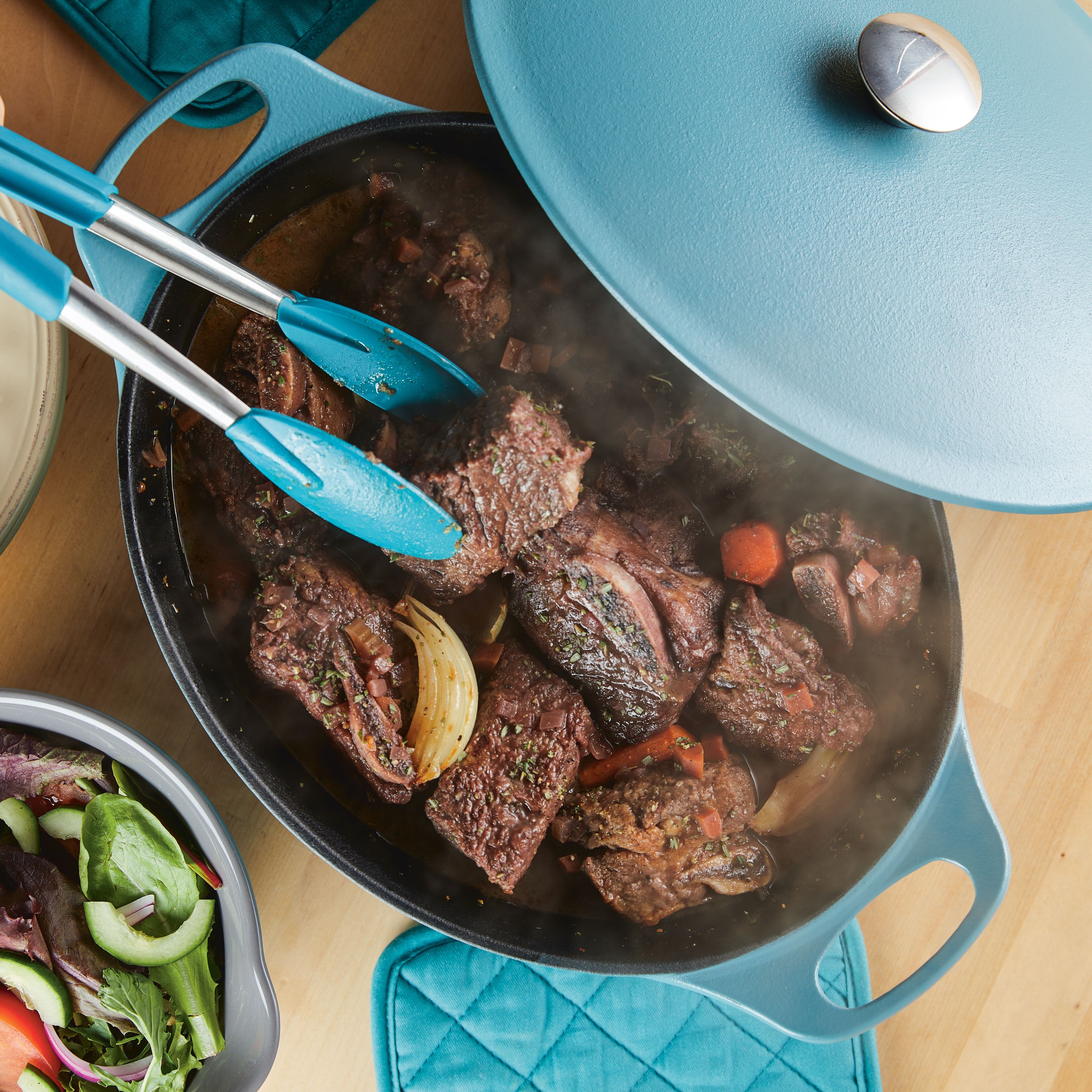 A close-up of an agave blue Rachael Ray NITRO Cast Iron 6.5-Quart Dutch Oven with the lid propped open, filled with a hearty beef stew with carrots and onions, and blue-tipped tongs resting on the side.