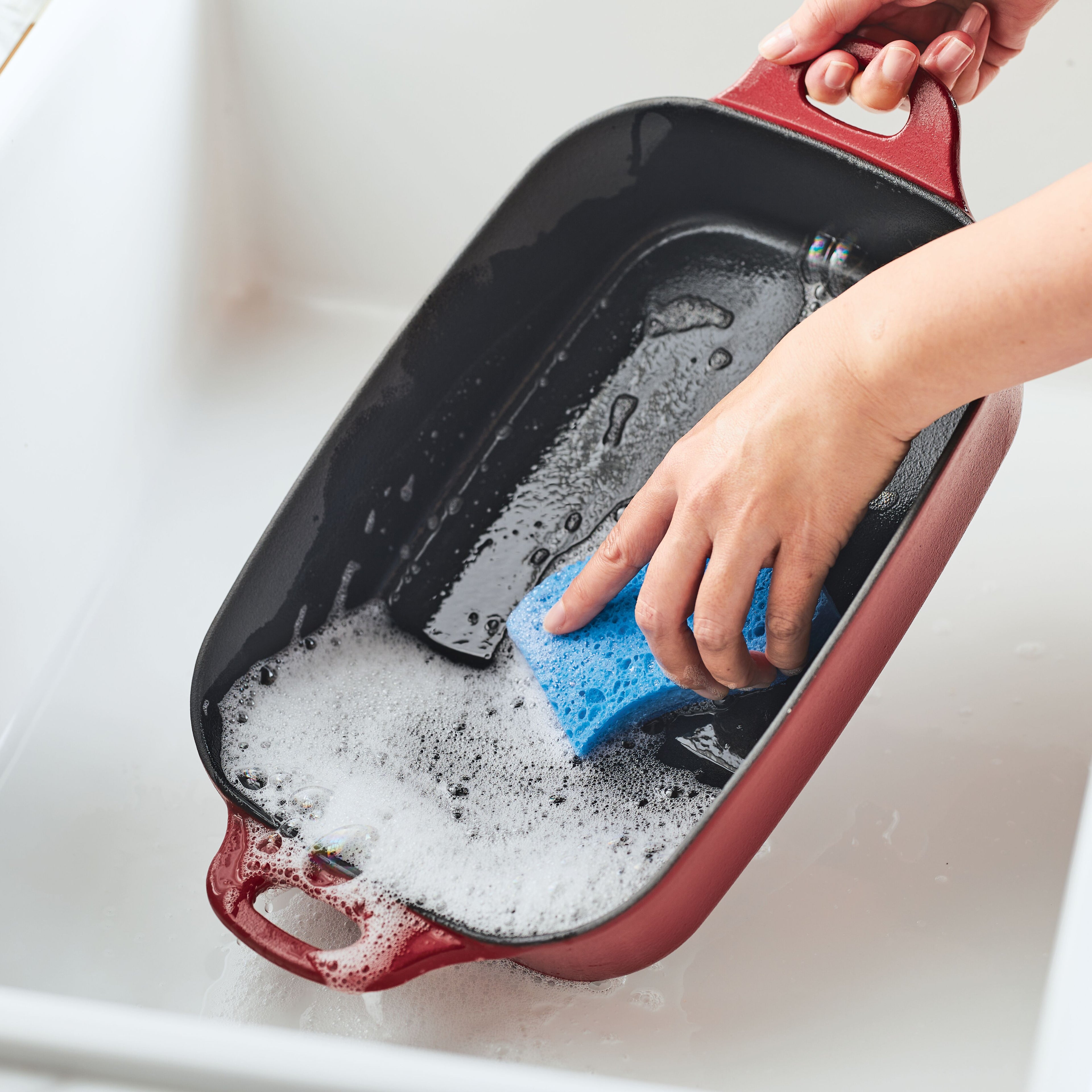 A close-up of a red Rachael Ray NITRO Cast Iron 9-Inch x 13-Inch Roasting Pan being cleaned in a white sink with soapy water and a blue sponge.