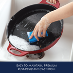 Hand washing a red cast iron pan with a blue sponge in a sink.