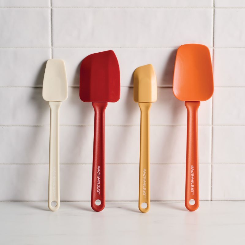 Four Rachael Ray silicone spatulas in various shapes and sizes are leaned against a white subway tile wall. The set includes a slim cream spatula, a large deep red spatula, a small yellow spatula, and a wide orange spatula, all featuring the Rachael Ray logo on the handles.