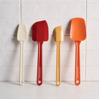 Four Rachael Ray silicone spatulas in various shapes and sizes are leaned against a white subway tile wall. The set includes a slim cream spatula, a large deep red spatula, a small yellow spatula, and a wide orange spatula, all featuring the Rachael Ray logo on the handles.