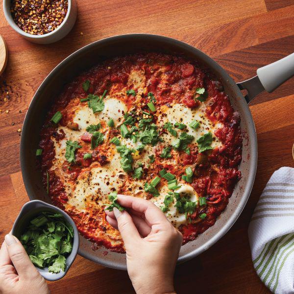 A person garnishes shakshuka with cilantro in a Cook + Create Nonstick Frying Pan, resting on a wooden table.