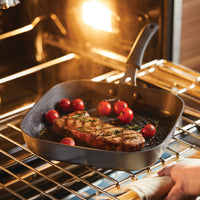 Steak and tomatoes sizzle in the 11-Inch Cook + Create Hard Anodized Nonstick Deep Grill Pan nestled inside the oven, ensuring even cooking with its exceptional scratch resistance.