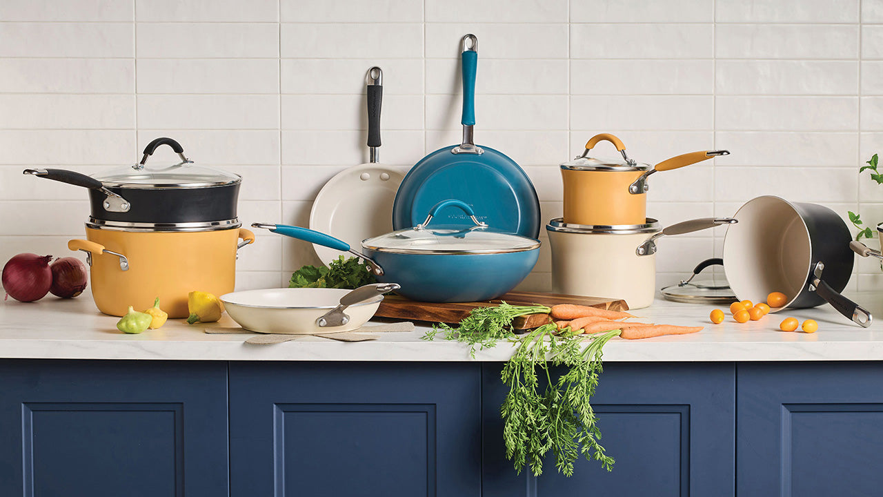 An assortment of Rachael Ray Cucina Ceramic cookware arranged on a white marble countertop against a white subway tile backsplash. The display includes a large stockpot, several saucepans, and skillets in a coordinated palette of agave blue, peppercorn gray, and almond, accented by fresh carrots and small yellow peppers.