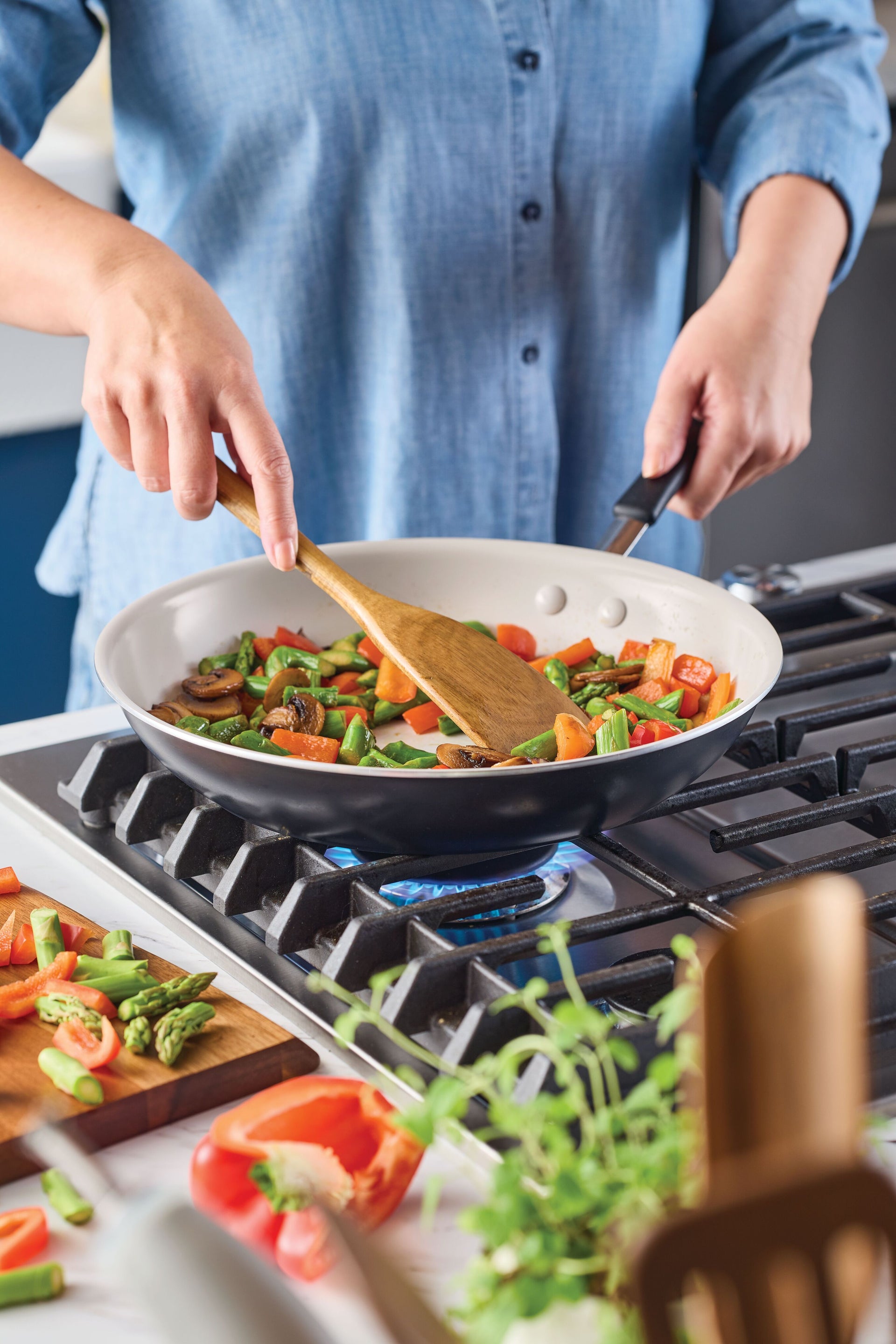 A person in a blue denim shirt using a wooden spatula to sauté vegetables in a Peppercorn-colored Rachael Ray Cucina Ceramic fry pan on a gas stovetop.