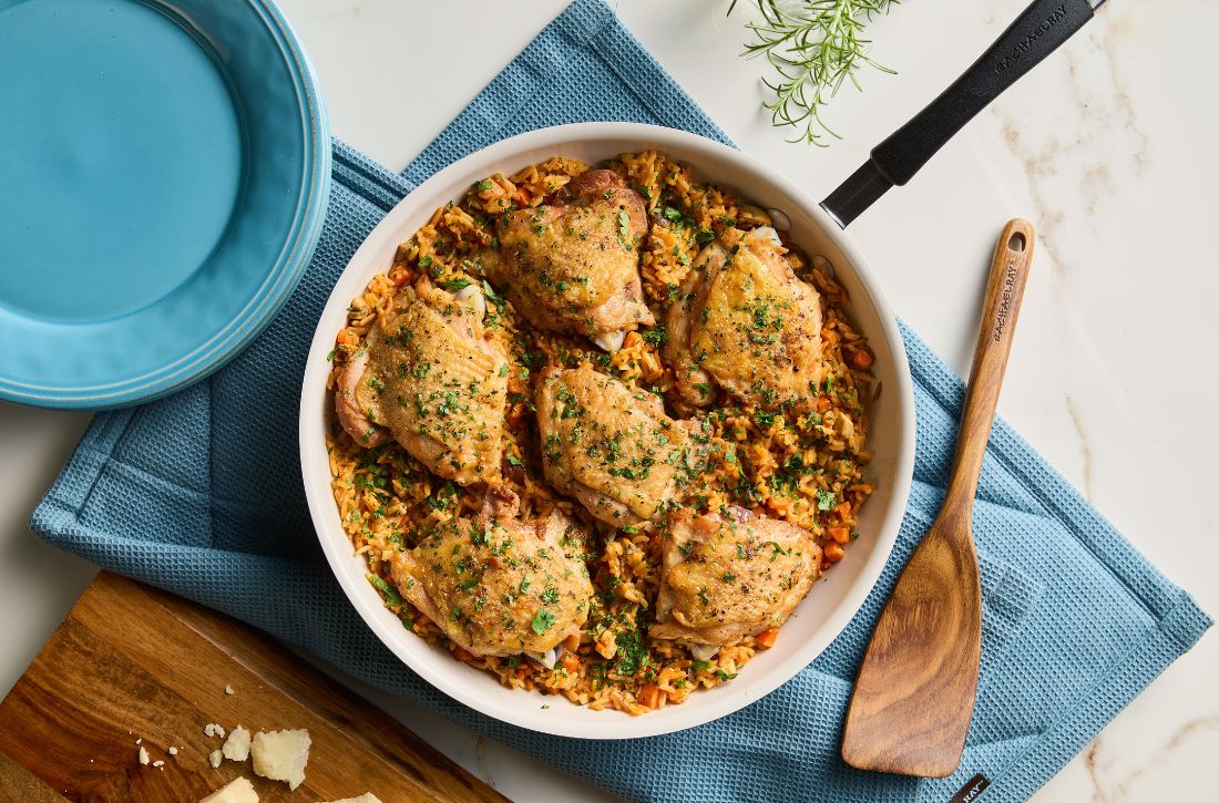 Italian-style chicken thighs and rice served in a Rachael Ray Cucina skillet on a blue kitchen towel with a wooden spoon.